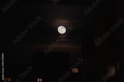 Atmospheric full moon shining over dark city residential buildings silhouettes in sleeping district at night under mysterious cloudy sky background