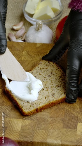 Hand using wooden tool to spread cheese on artisan loaf. Closeup of skilled hand smoothing cheese onto rustic bread among kitchen ingredients