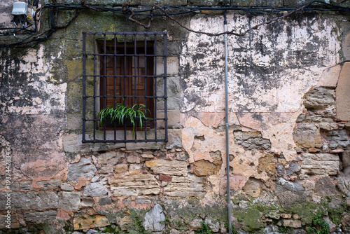 Old rustic stone wall with a small wooden window behind a black iron grate and a green plant in a pot on a weathered facade of a medieval building in a spanish village