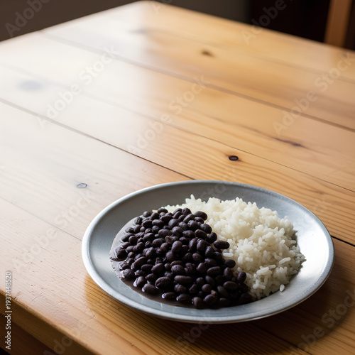 Simple serving of rice and black beans on a plain metal plate placed in the lower right corner of a large light wooden table, most of the table left empty as negative space.