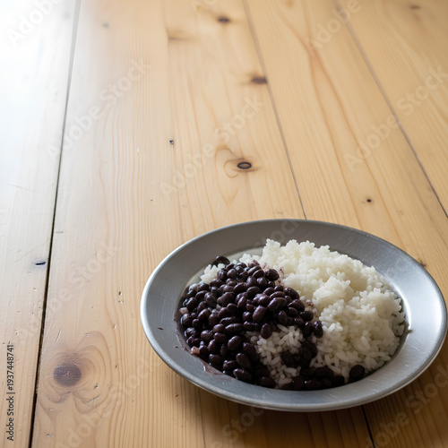 Simple serving of rice and black beans on a plain metal plate placed in the lower right corner of a large light wooden table, most of the table left empty as negative space.