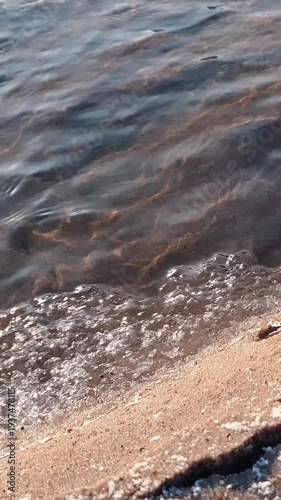 Vertical closeup shot of clean waves of shallow river or creek water surface and frozen snow-covered sand shore with ice crust at sunny winter or spring day. Aquatic texture pattern natural background
