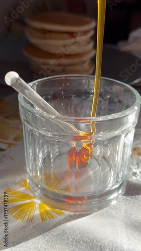 Close-up of honey being poured into a glass jar, with stacked pancakes in the background. Perfect for food content, breakfast cooking, and food styling videos.