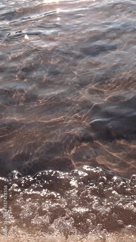 Vertical close up shot of clean flowing waves of shallow water stream surface with sand bottom and frozen ice-covered shore at sunny winter or spring day. Aquatic texture pattern natural background.