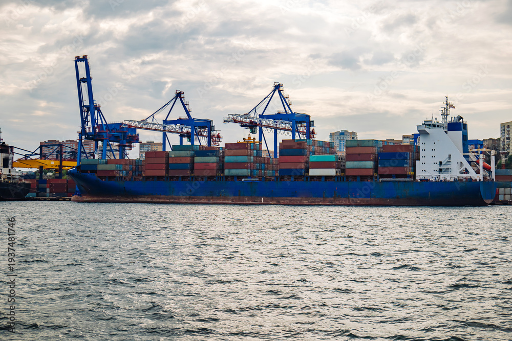 Fototapeta premium Cargo vessel docked at vladivostok port, stacked multicolored containers blocking cloudy maritime skyline