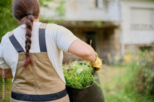 Female gardener with ponytail carries a heavy basket filled with grass clippings produced after mowing a garden during outdoor maintenance work.