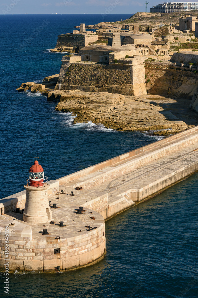 Fototapeta premium Faro de cúpula roja en el rompeolas de La Valeta con Fort Ricasoli al fondo. Arquitectura histórica de defensa militar y navegación en el Mar Mediterráneo.