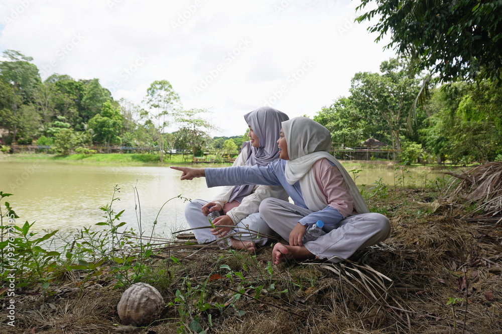 Fototapeta premium Two happy young Asian Muslim sisters sitting under a tree by the lake at sunset, chatting and laughing together