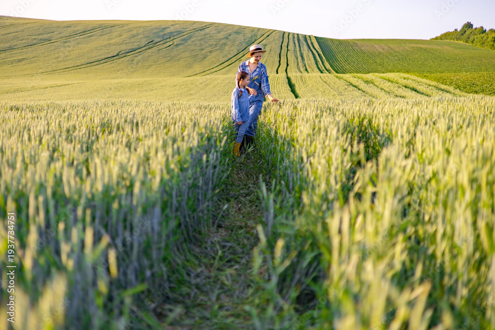 Fototapeta premium A woman and a young girl walk through a lush green wheat field under a clear sky, showcasing agricultural beauty and family connection