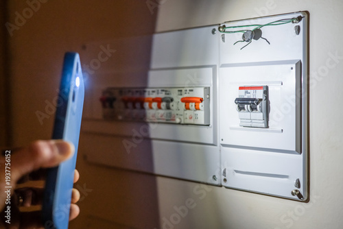 Person holding smartphone flashlight to illuminate a tripped electrical fuse box during a home power outage, highlighting emergency troubleshooting and domestic electrical issues