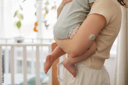 Close-up of a woman in light homewear with a Continuous Glucose Monitoring on her arm holding a baby next to a white crib with a hanging musical mobile toy. 