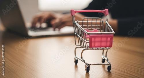 Small empty shopping cart on wooden table with blurred person working on laptop in background suitable for retail shopping ecommerce online store advertising