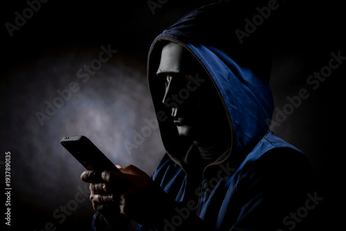 A scammer in a dark room, hiding his face under a black mask, uses a phone to deceive people.