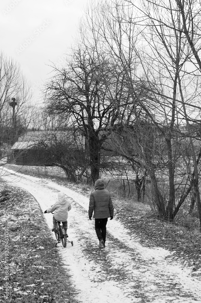 Obraz premium Walking together on a snowy path as a child rides their bike near bare trees in a tranquil winter landscape in a quiet rural setting during the early afternoon