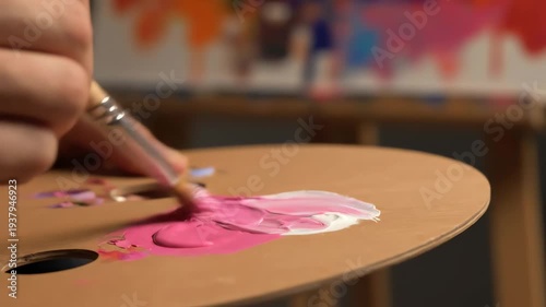 Close Up of Paintbrush Mixing Pink and White Paint on Palette in Studio Setting With Colorful Abstract Painting in Background