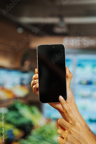 Hand holding smartphone with blank screen in grocery store, finger tapping display. Blurred supermarket produce and refrigeration background, concept for shopping app, mobile payment, scanning.