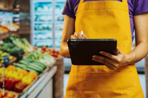 Grocery store worker in yellow apron using a tablet for inventory in the produce aisle, with fresh vegetables blurred in the background.