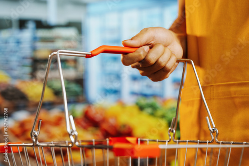 Fotografie Close-up of shopper hand holding red handle of metal grocery basket in supermarket, colorful blurred produce aisle background, retail shopping and consumer lifestyle concept