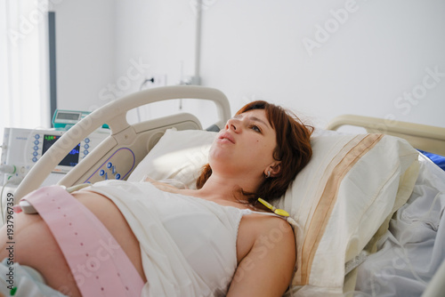 Tablou pe pânză Pregnant woman lying in a hospital bed with a fetal monitoring belt, looking anx