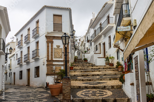 One of the picturesque streets of Frigiliana, a white Andalusian village, with whitewashed facades decorated with potted plants and flowers, and traditional mosaic floors. Málaga, Spain