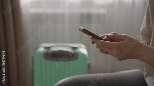 Close up of female hands using a smartphone to book a taxi online, preparing for departure with a suitcase in the background, symbolizing travel and modern transportation booking.