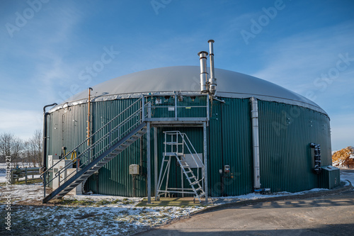 Photography Exterior view of agricultural biogas plant with large anaerobic digester tank and domed membrane roof equipped with external piping, ventilation stacks and access platform with stairs for maintenance