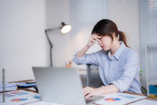Stressed asian female worker sitting at office desk, experiencing headache and burnout from overwork, holding hand to forehead while looking at laptop