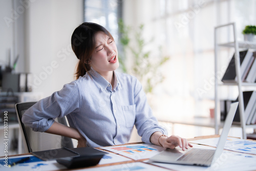 Asian business woman feeling lower back pain during long hours of work, experiencing discomfort and stress from her office job, needing physical relief