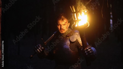 A knight in full armor, his face bloodied and scarred, holding a sword and torch, is sneaking through a dark night forest. A medieval warrior searches in the darkness.