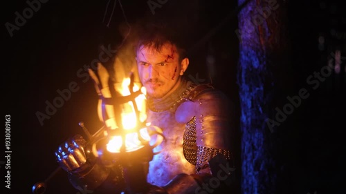 A knight in full armor, his face bloodied and scarred, holding a sword and torch, is sneaking through a dark night forest. A medieval warrior searches in the darkness.