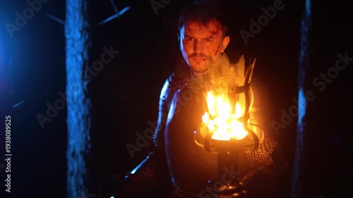 A knight in full armor, his face bloodied and scarred, holding a sword and torch, is sneaking through a dark night forest. A medieval warrior searches in the darkness.