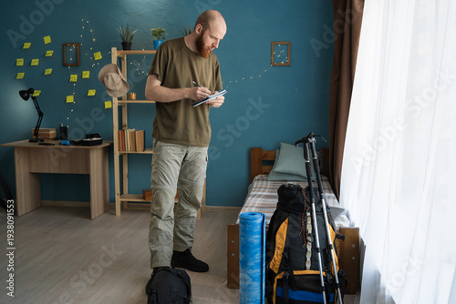 active traveler making checklist for mountain journey standing in home interior. Hiker preparing for hiking trip and trekking gear organization.