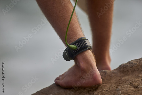 Close-up of a surfer’s ankle secured with a safety leash while stepping toward the ocean, capturing the anticipation of entering the waves. The image highlights the connection between surfer and board
