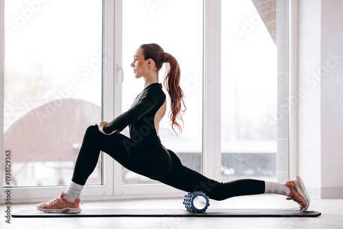 Young woman stretching with foam roller during a low lunge home fitness workout by the window