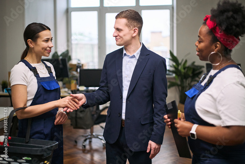 Caucasian young adult man shaking hands with Caucasian young adult woman in cleaning uniform, while Black young adult woman in cleaning uniform holding clipboard observing in office setting