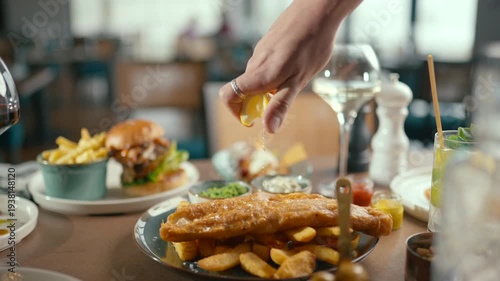 Hand squeezes lemon over golden fried fish and chips. Restaurant table with burgers and drinks nearby. Warm lighting, casual dining atmosphere. Great for food ads or restaurant promotions