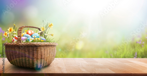 A wicker basket filled with painted easter eggs, grass and daffodils sat on a wooden table in a meadow to celebrate a religious event, Easter, with a wide banner style display background.