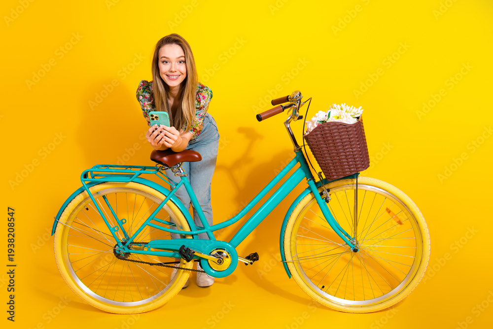 Naklejka premium Young chic woman with turquoise bicycle and basket of flowers smiles against a bright yellow background