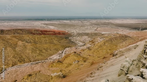 Aerial view of a vast desert landscape with layered rock formations and dry terrain. The scene features a mix of earthy colors and distant mountains.