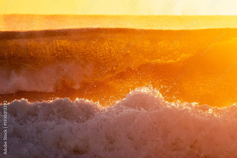 Fototapeta premium Waves crash onto the shore during sunrise, creating a dynamic scene with sea spray and mist. The golden light of the rising sun illuminates the ocean, giving the spray a vibrant glow.