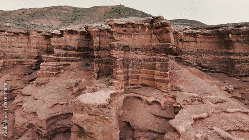 Aerial view of a rugged canyon with layered rock formations and sandy terrain. The landscape features earthy tones and steep cliffs.