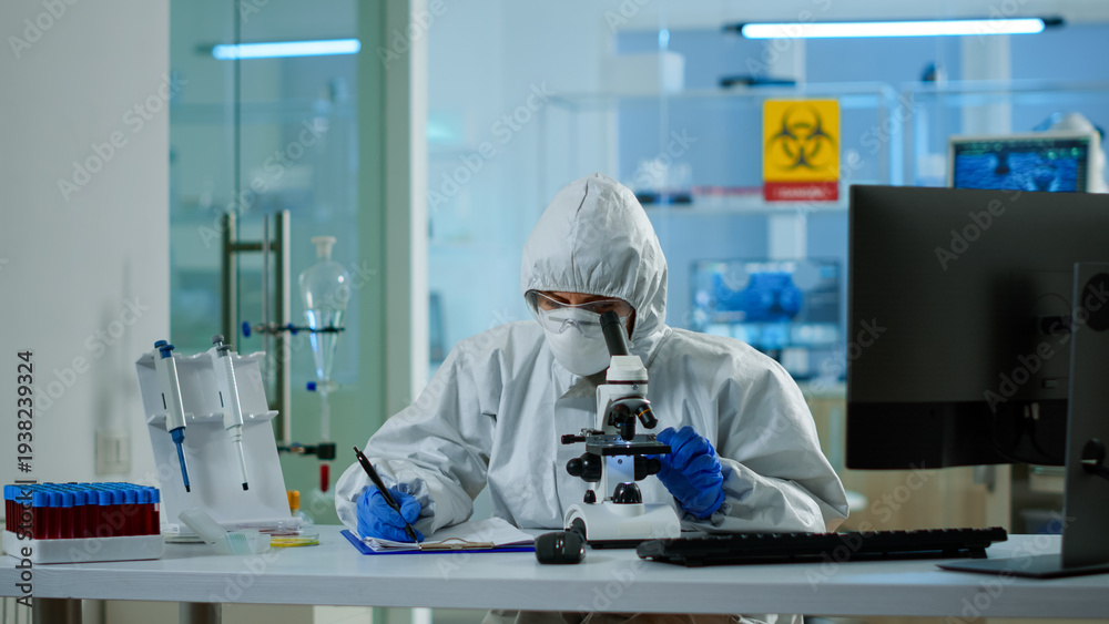 Naklejka premium Biologist in ppe suit working on microscope and writing information on clipboard in equipped laboratory. Chemist examining vaccine evolution using high tech researching diagnosis against covid19 virus