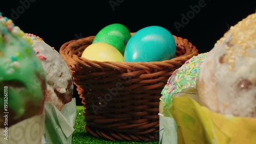 Closeup of easter turquoise and yellow eggs in wicker basket and glazed cakes with colorful sprinkles on soft icing. Spring composition on green grass and dark background. Macro of festive symbols