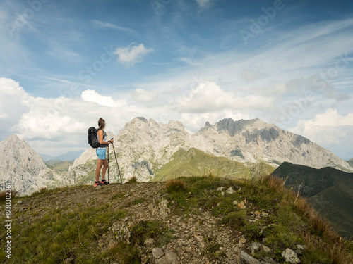 A woman with a backpack and hiking poles stands on a grassy mountain ridge, looking at the mountains in the distance, the Coglians gr under a blue sky. Monte Crostis,Carnia,Friuli Venezia Giulia,Italy