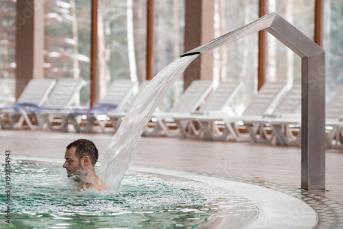 A man in a pool under a waterfall for a back and neck massage, with sun loungers in the background. High-pressure water supply. Concept of relaxation, spa treatments, and rejuvenation in a hotel. 
