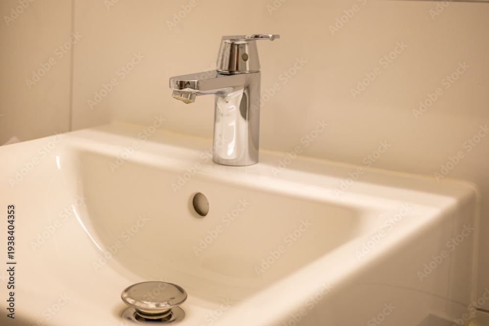 Fototapeta premium Modern white ceramic bathroom sink with chrome mixer tap, clean hotel bathroom interior, hygiene and sanitation concept, overflow hole visible, drain plug, cozy home interior, morning hygiene