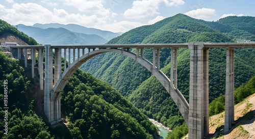 A majestic stone arch bridge spans a turquoise mountain river, blending historic architecture with the rugged landscape of a nature tourism landmark under a clear blue sky