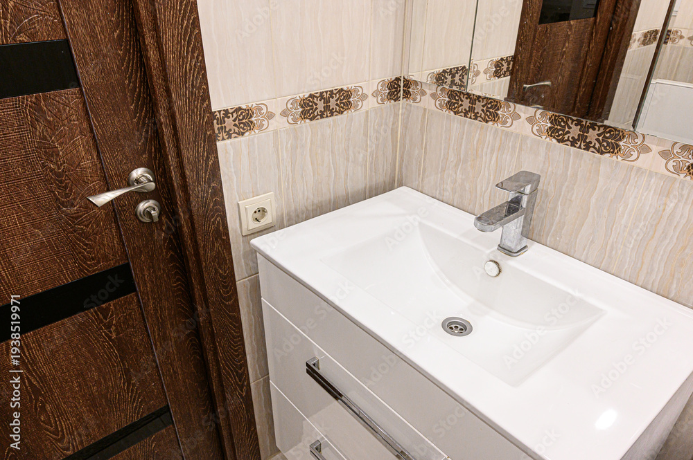 Fototapeta premium sleek white bathroom sink with a modern chrome faucet sits against beige tiled walls with decorative border, beside a dark wood door with metallic handle.
