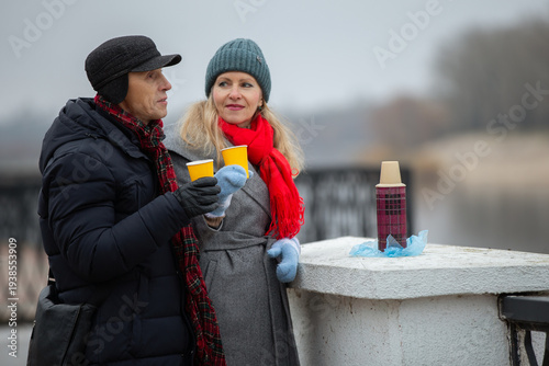 Couple enjoys warm drinks by the river in winter, wrapped in cozy clothes and sharing a moment of joy