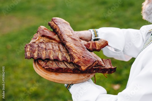 Smoked pork ribs on a wooden platter held outdoors by a person in traditional embroidered shirt